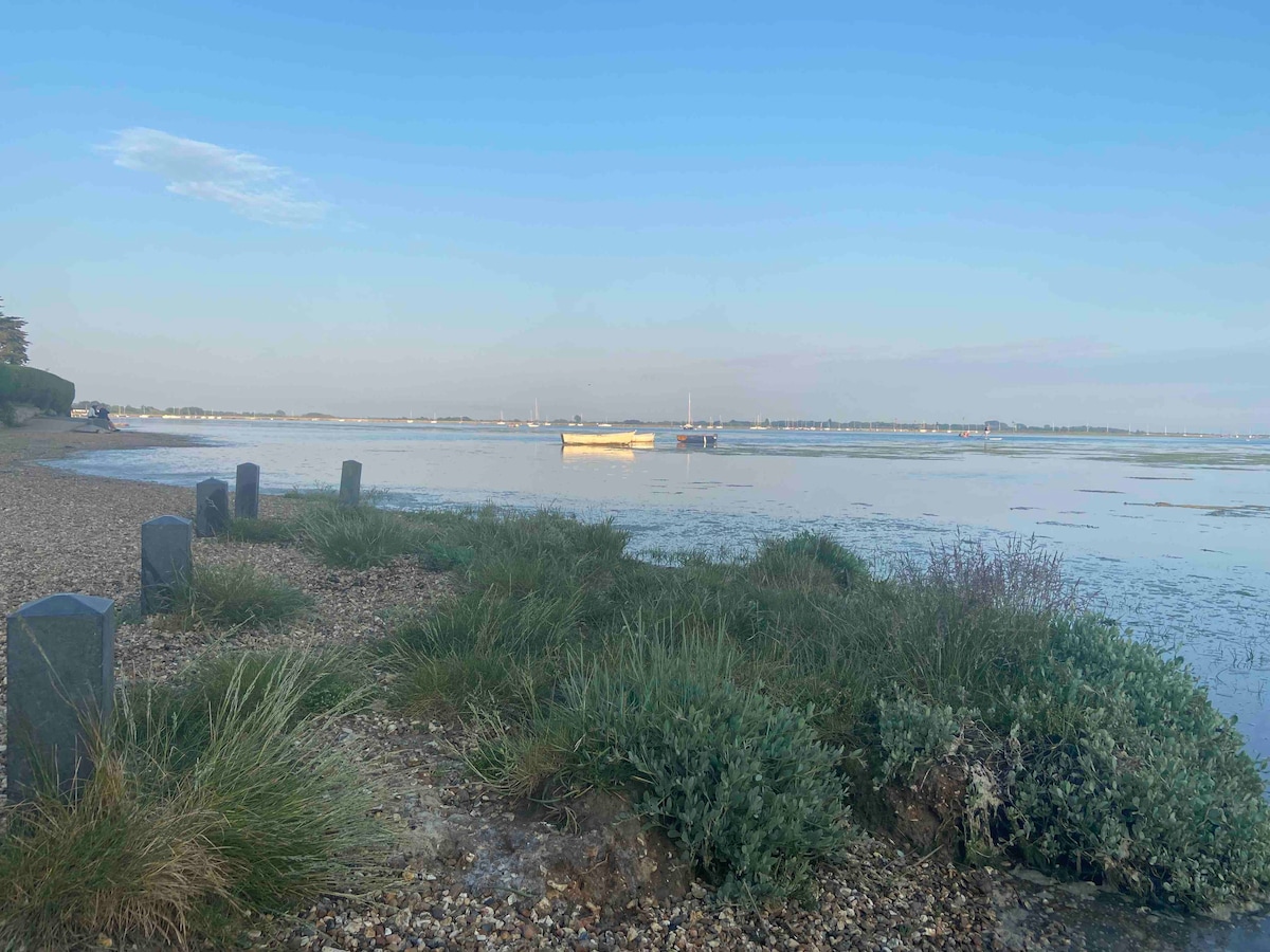 A serene view of a calm waterway is depicted, featuring a single boat gently bobbing in the distance. Lush green grasses and pebbles line the shore, adding natural texture to the scene. The sky is clear, reflecting soft blue hues over the water.