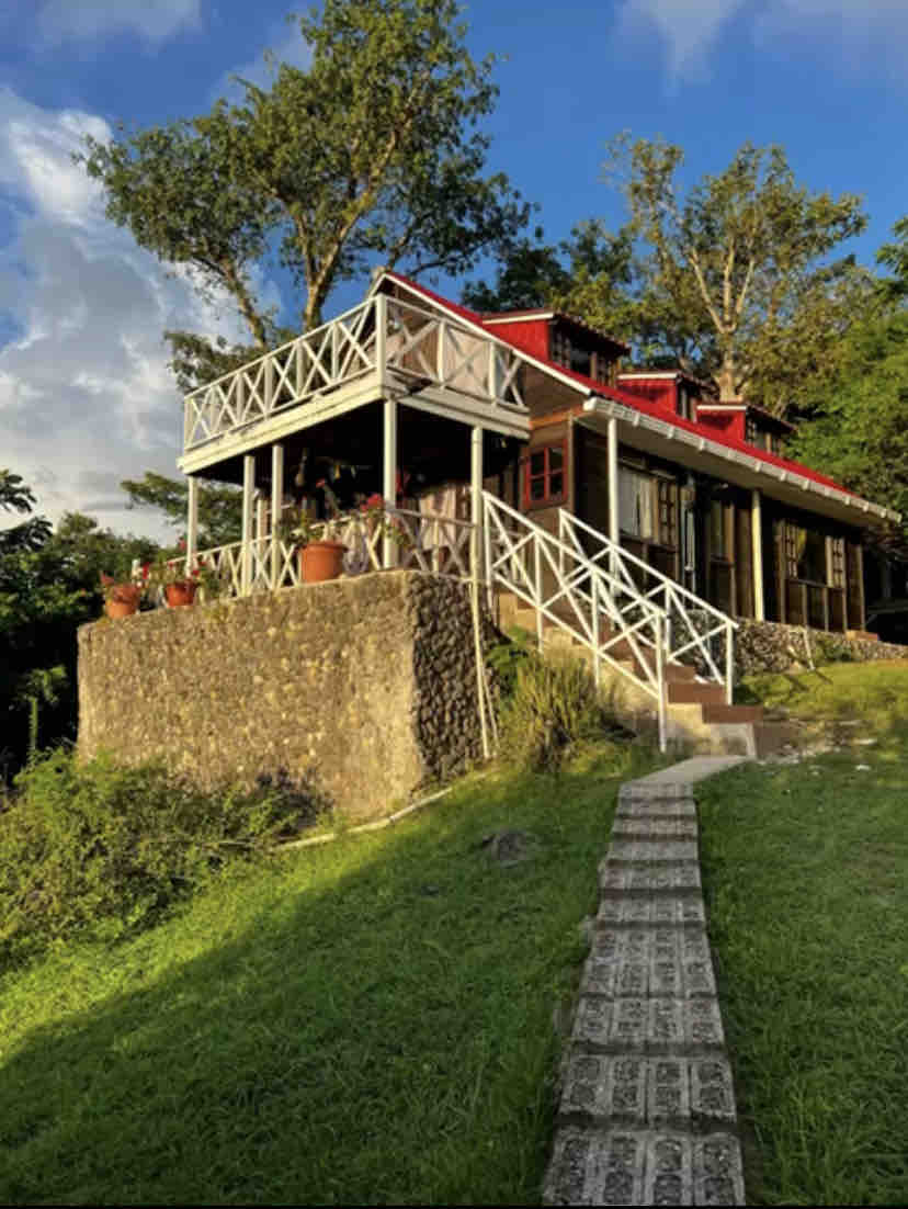 The two-story house is nestled on a hillside, featuring a spacious terrace with a railing and potted plants. Stairs lead up to the entrance, while a pathway of stone tiles meanders through the lush greenery surrounding the property.