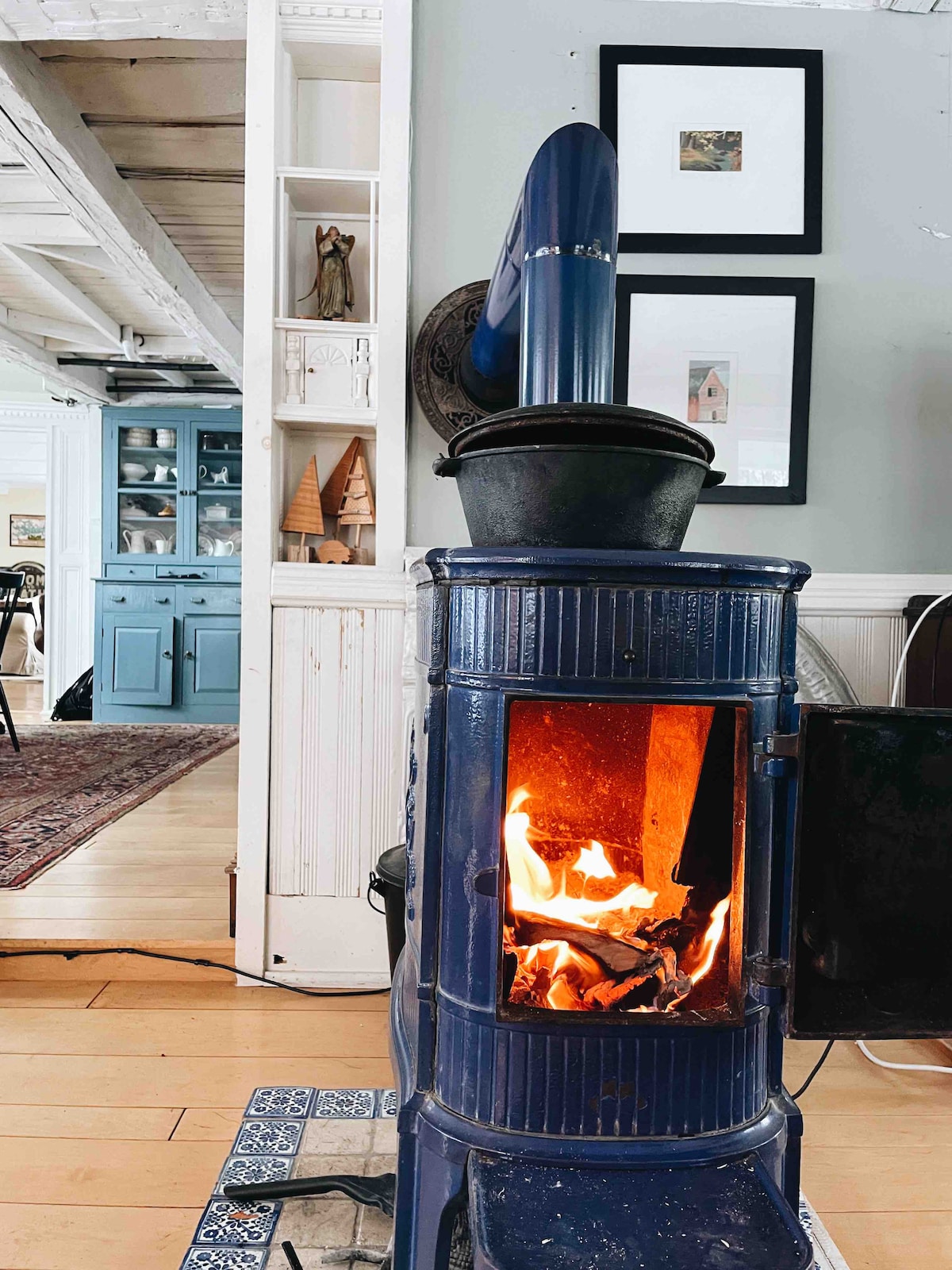 A blue wood-burning stove is showcased with flames dancing inside. The warm glow from the fire illuminates its surroundings, including a white wall adorned with framed artwork. A glimpse of a blue kitchen cabinet can be seen in the background.