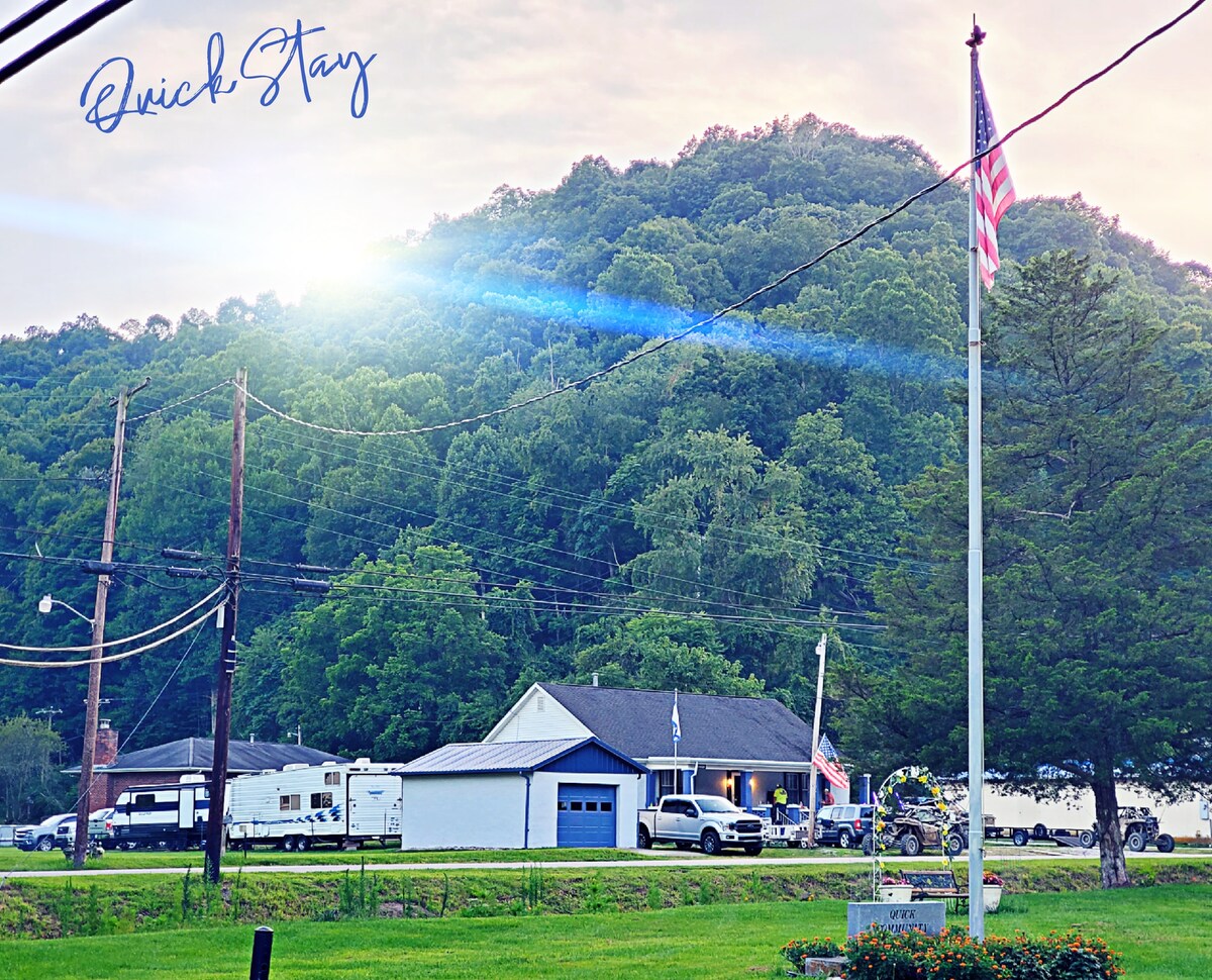 The image shows a classic WV house set against a backdrop of lush green mountains. A flagpole with an American flag stands in the foreground. Nearby, a trailer is parked beside the house, with multiple vehicles in the driveway, indicating parking accommodations.