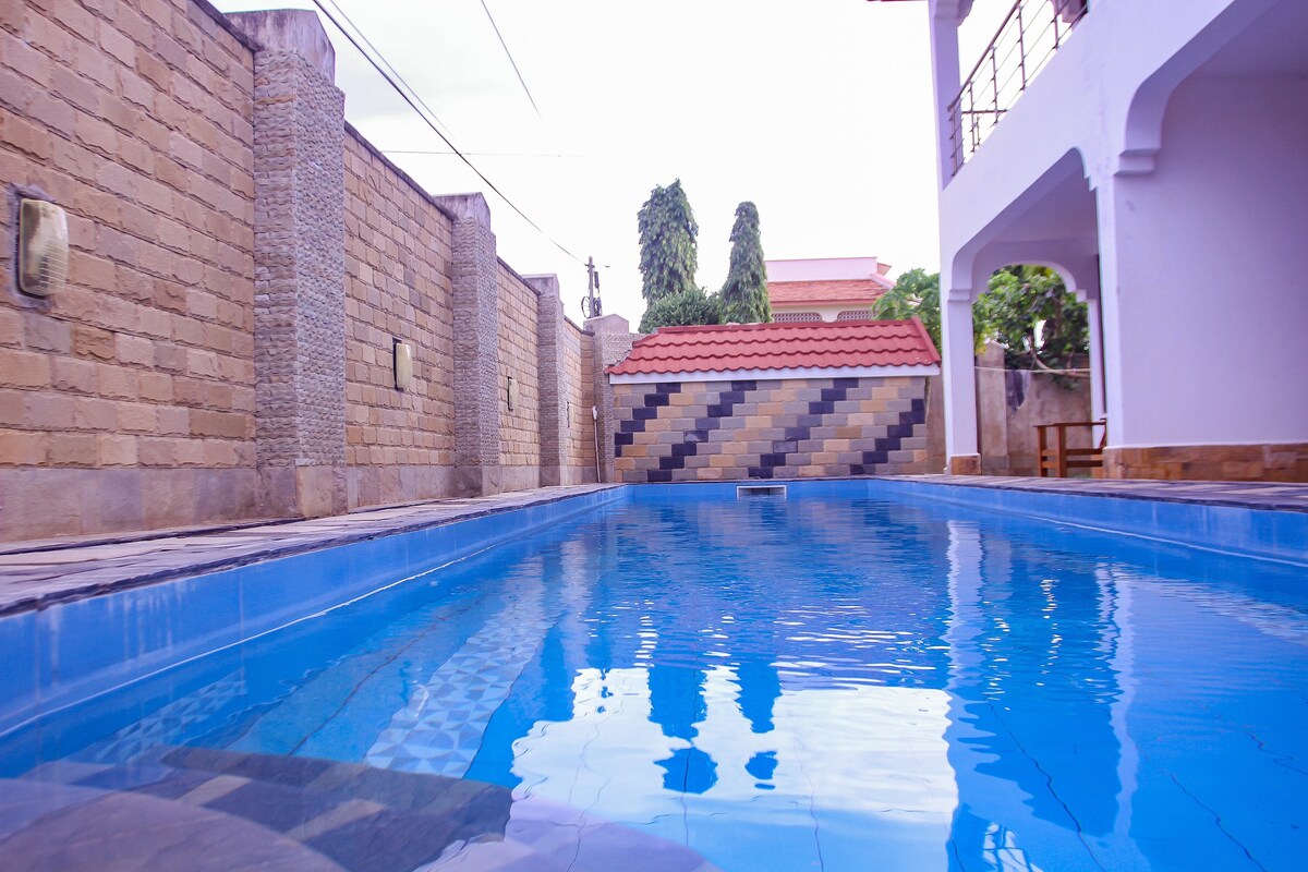 A sparkling blue swimming pool is visible, surrounded by textured stone walls and a well-maintained patio area. The water reflects the clear sky and nearby greenery, providing a tranquil space for relaxation. The backdrop includes a decorative wall featuring a patterned design.