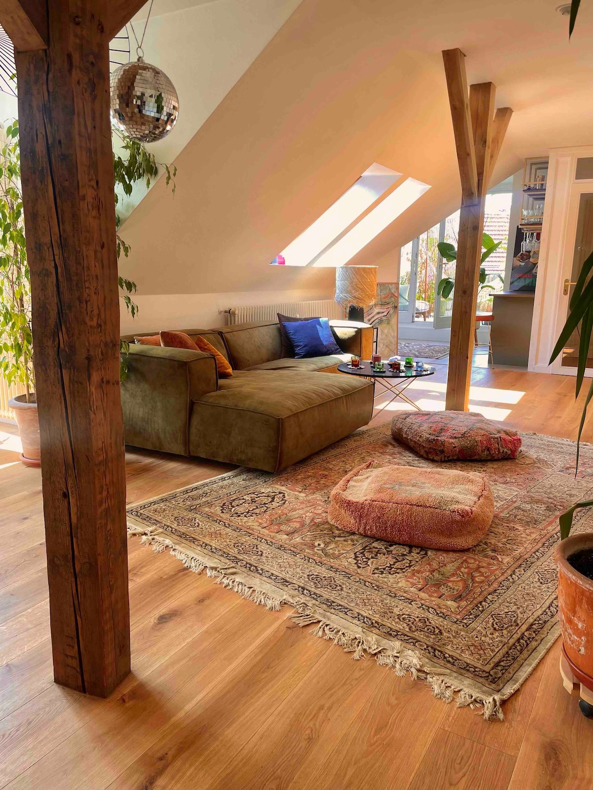 A cozy living area is highlighted by a large, comfortable couch and vibrant cushions on a decorative area rug. Natural light floods through the skylight and nearby windows, creating a warm ambiance. Decorative plants and wooden beams add character to the space.