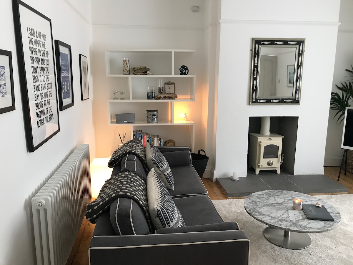 A spacious living area features a comfortable gray sofa adorned with patterned cushions. A round marble coffee table is placed centrally, and a decorative mirror is mounted on the wall. Soft lighting and a stylish log burner contribute to a cozy ambiance.