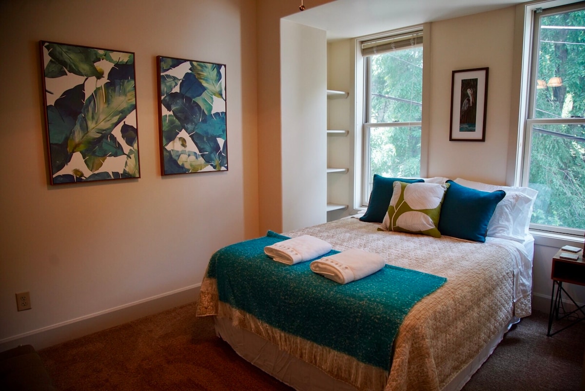 A bright and inviting bedroom features a spacious bed adorned with a textured blanket and decorative pillows. Two artworks with leaf motifs are hung on the wall, while natural light filters in through two large windows.