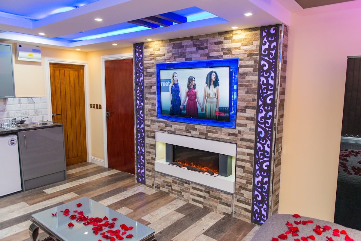 An open-plan living area features a modern electric fireplace beneath a large flat-screen TV, set against a stone feature wall. Soft lighting and decorative rose petals enhance the inviting ambiance. A sleek kitchenette is visible in the background, along with tasteful furnishings.