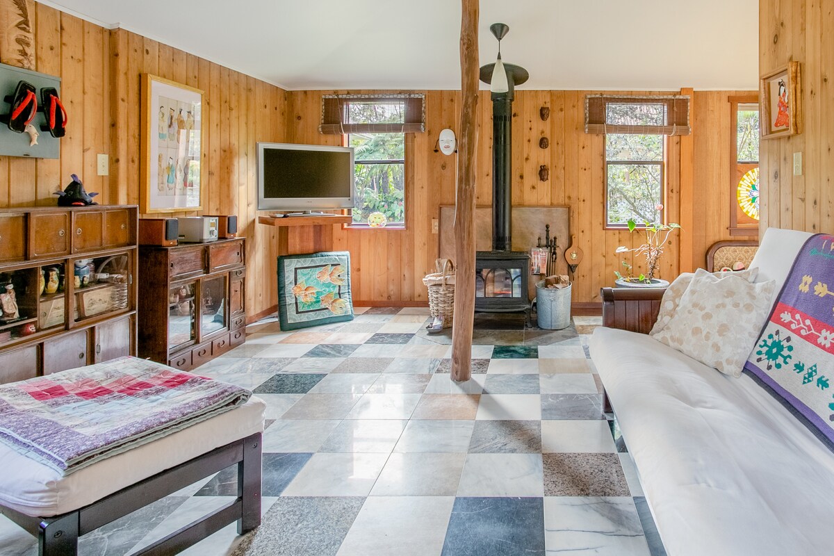 The living room features a combination of wooden walls and a patterned tile floor, creating a warm atmosphere. A wood-burning stove serves as a focal point, while windows allow natural light to illuminate the space. A comfortable seating area is present, enhancing the cozy setting.