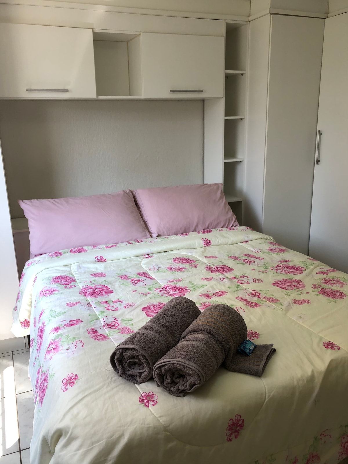 A neatly arranged sleeping area features a cozy bed with pink pillows and a floral quilt. Two rolled towels are placed at the foot of the bed. Light-colored built-in cabinetry and shelves provide ample storage space, enhancing the room's functionality.