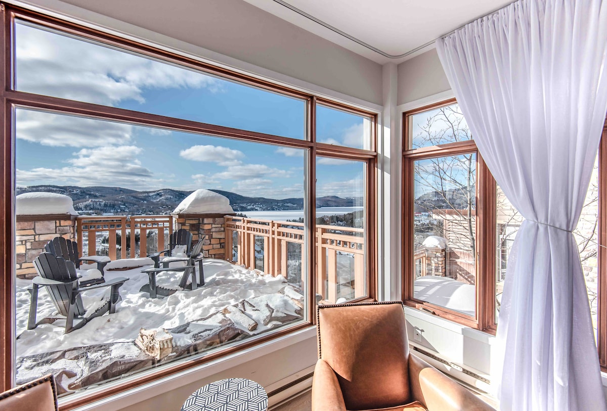 A large window offers expansive views of the snow-covered landscape and distant mountains. Two outdoor chairs are positioned on a terrace surrounded by snow, providing a serene spot for relaxation. Soft natural light enhances the warm tones of the cozy seating area inside.