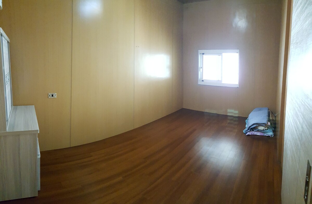 The spacious room features smooth wooden flooring and light-colored walls. A single window allows natural light to enter, illuminating the uncluttered space. A neatly stacked arrangement of blankets is visible on the floor, and a simple dresser is positioned against one wall.