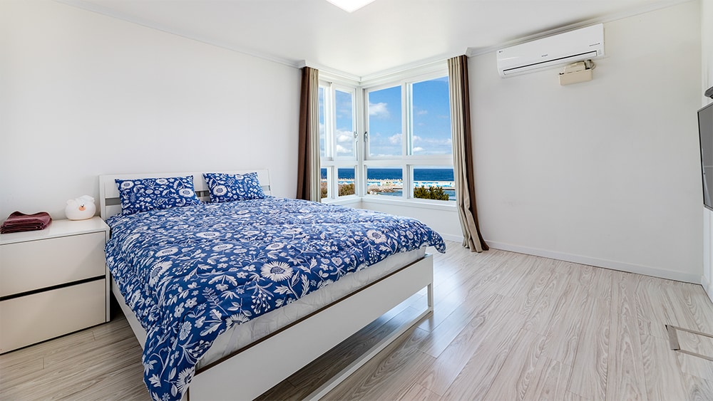 A bright bedroom features a spacious bed with a blue floral duvet cover. Large windows provide expansive views of the sea, while natural light enhances the airy atmosphere. An air conditioner is mounted on the wall, and a bedside table is positioned next to the bed.