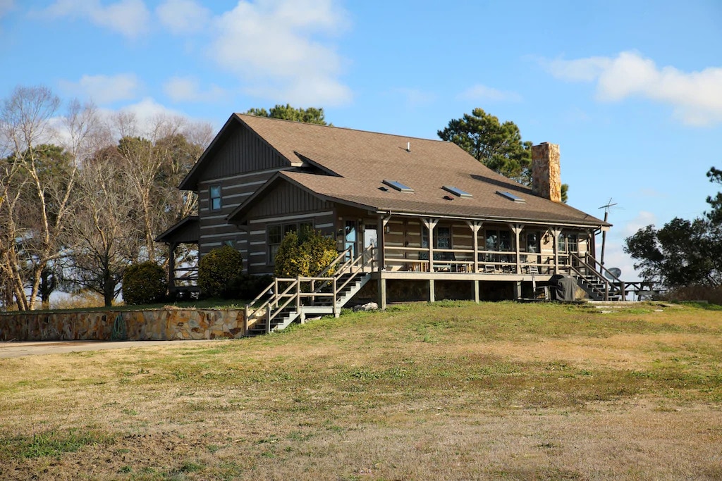 The hunting lodge features a two-story design with a large front deck and stone accents. Expansive windows provide views of the surrounding landscape. The grassy area surrounding the lodge is complemented by nearby trees and a gentle slope.
