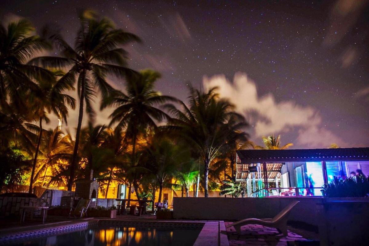 A serene nighttime scene is captured, featuring swaying palm trees against a starry sky. The warm glow from the nearby home illuminates the outdoor area, and reflections shimmer on the surface of the pool.