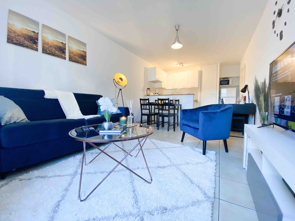 A modern living area showcases a blue sofa and two blue armchairs, arranged around a glass coffee table. A plush area rug covers the tiled floor. The space is illuminated by a ceiling light and features a dining table set for four in the background, with a fully equipped kitchen visible.