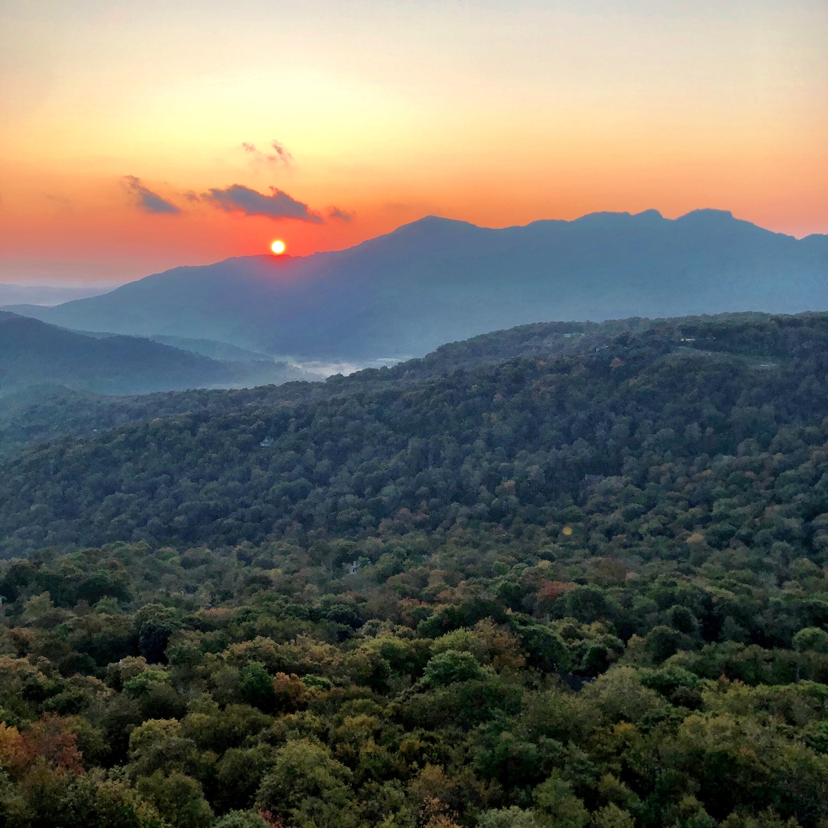 A stunning view of the sun setting behind majestic mountains, casting a warm glow over the lush, green landscape. The horizon is painted in soft oranges and yellows, showcasing the beauty of the surrounding Blue Ridge Mountains.