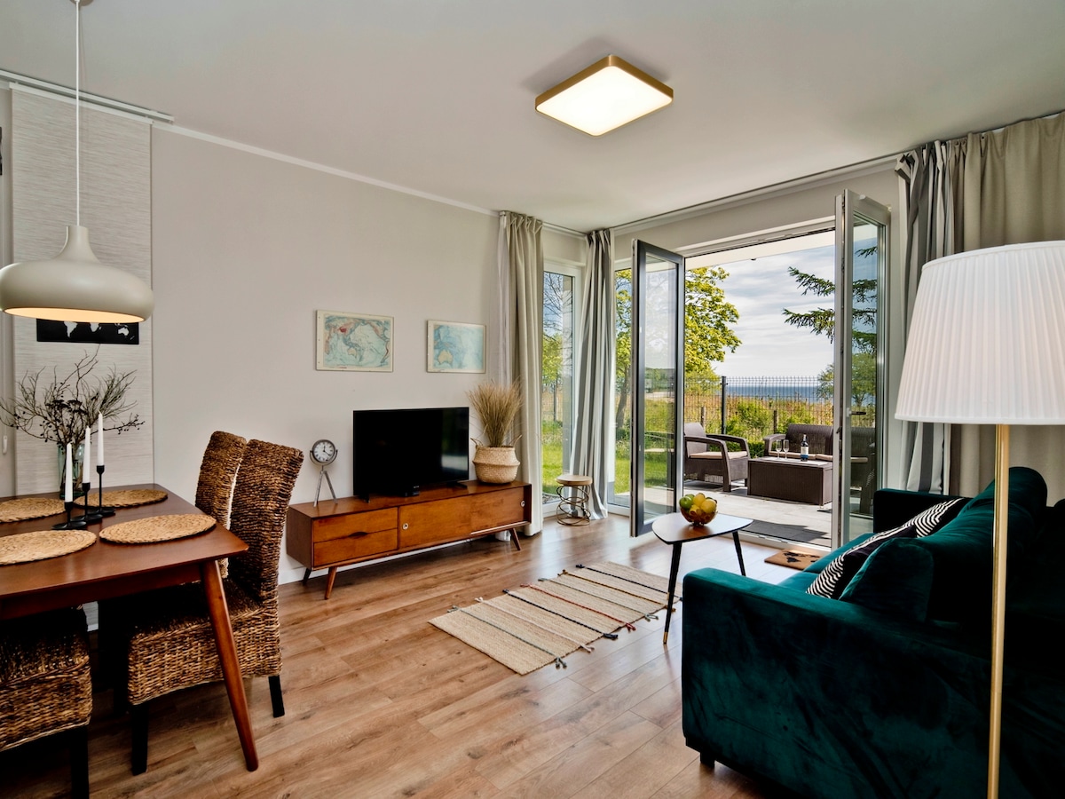 A living area features a dining table with woven chairs, a television on a wooden console, and a plush green sofa. Large glass doors open to a terrace with garden views, allowing natural light to fill the space.