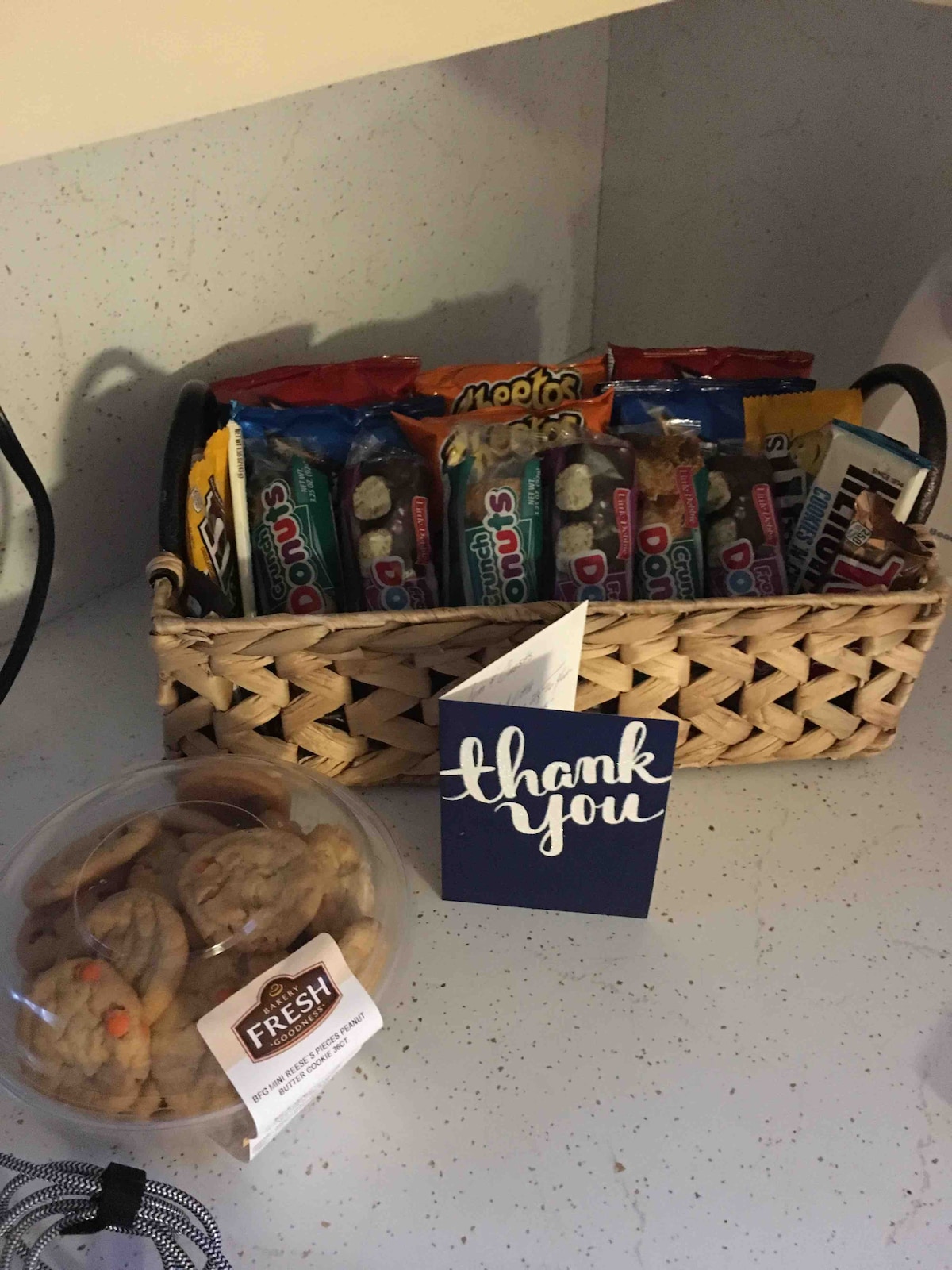 A woven basket is filled with various snacks, including candy bars and granola bars. A container of freshly baked cookies sits next to the basket. A thank-you card is propped against the basket, offering a warm welcome to guests.