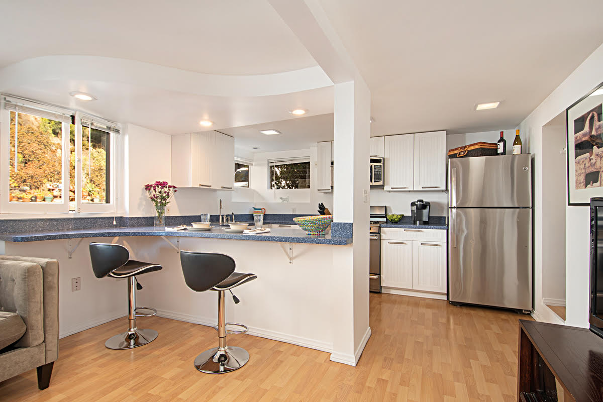 A modern kitchen area is showcased with white cabinetry and stainless steel appliances. Two stylish bar stools are positioned at a blue-topped counter. Natural light fills the space through multiple windows that provide views of greenery outside.