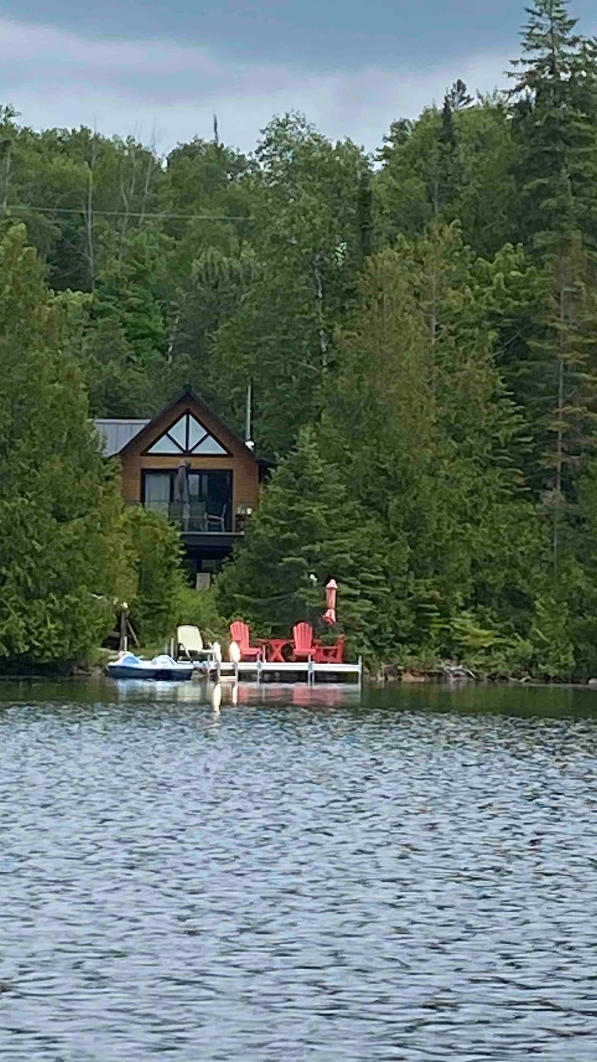 A tranquil view of a chalet surrounded by lush greenery, set on the edge of a calm lake. A private dock featuring seating and watercraft is visible in the foreground, reflecting the peaceful environment and natural beauty of the area.