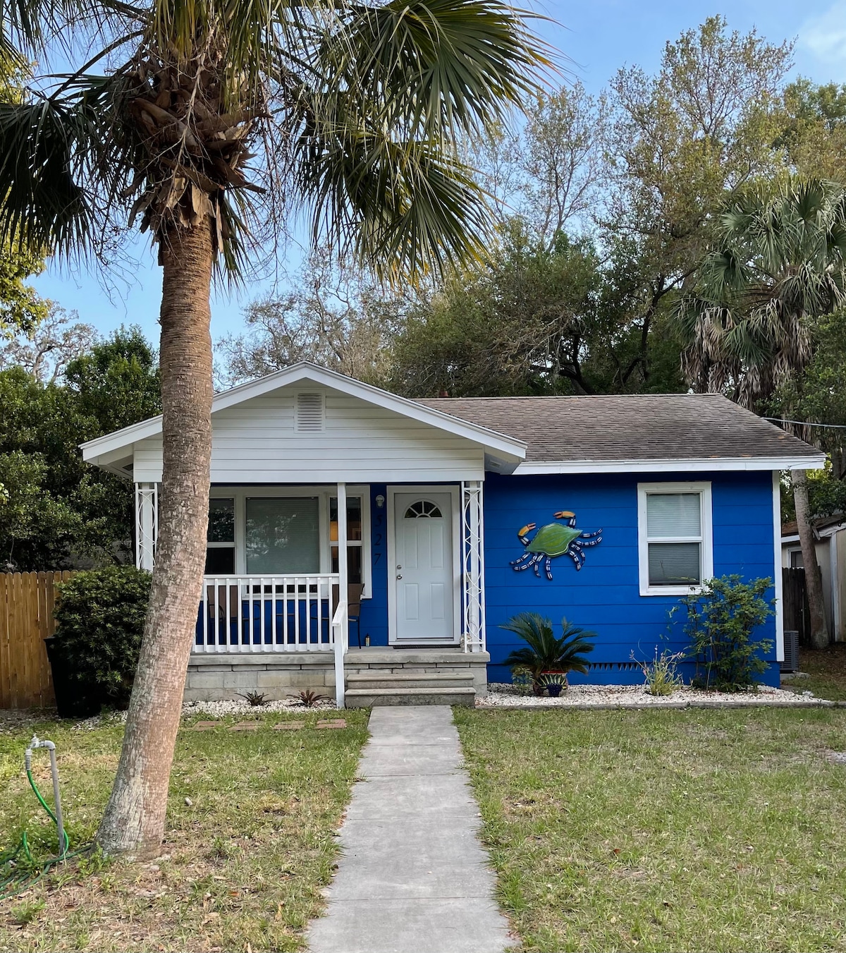 A charming blue house is seen, featuring a white porch and inviting front door. A playful mural of a crab adorns the exterior wall, while a shaded front yard includes lush greenery and palm trees. A concrete pathway leads to the entrance.