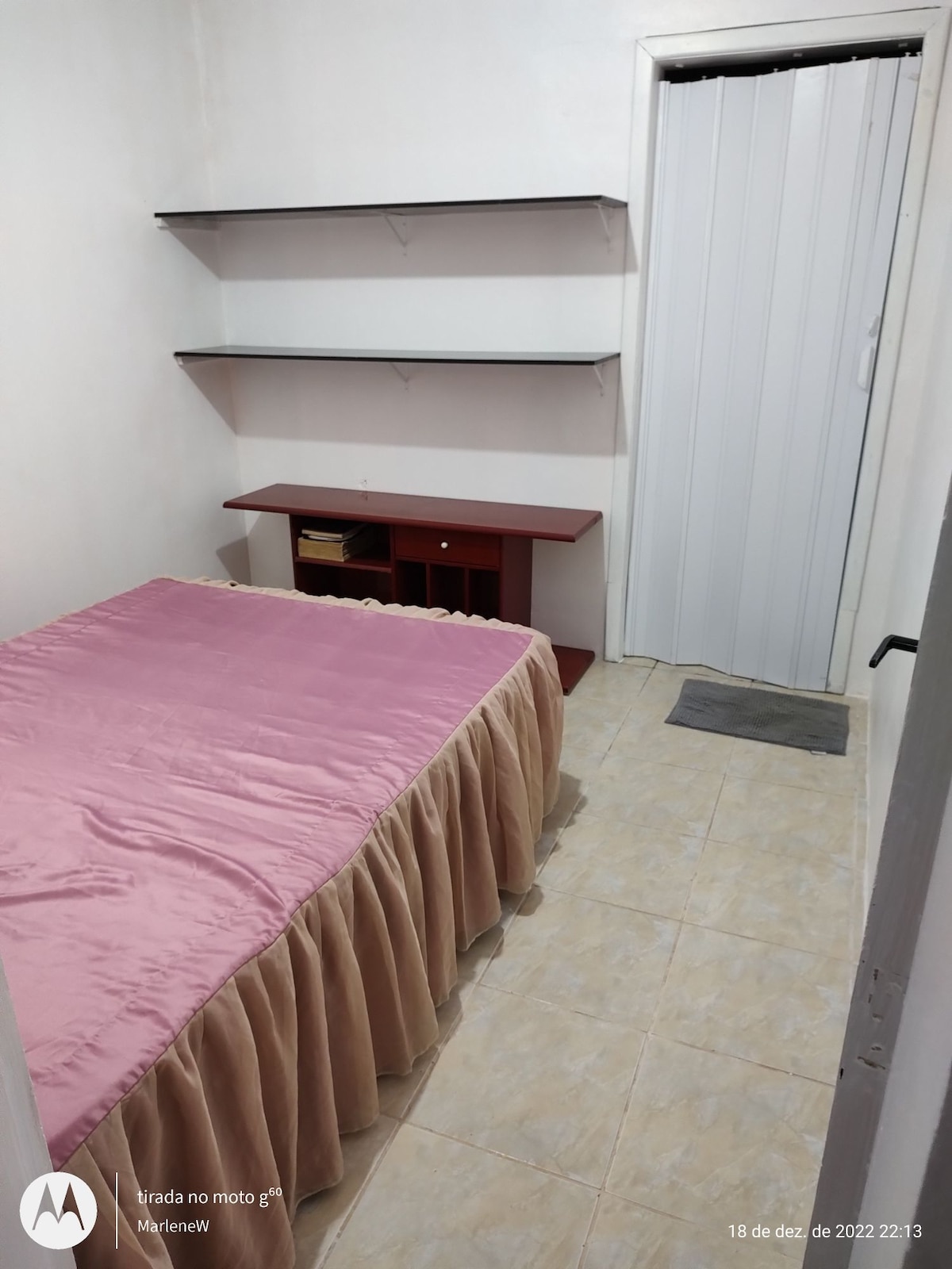 A simple bedroom is presented with a spacious bed dressed in a light pink bedspread. A wooden shelf is mounted on the wall, providing additional storage options. The room features tiled flooring and a doorway leading to another area, with a small mat positioned at the entrance.