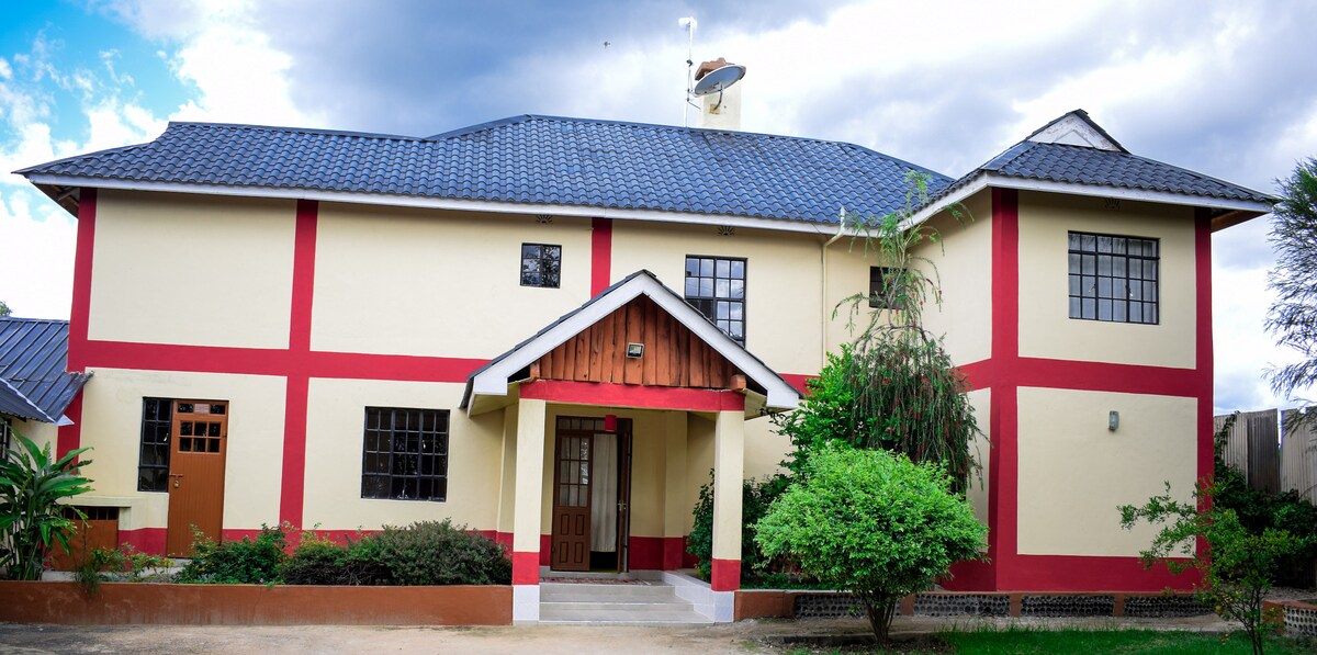 The exterior of a two-story house is shown, featuring a combination of cream-colored walls with red accents. A central gabled entrance is flanked by two large windows. The surrounding landscape includes manicured shrubs and plants under a partly cloudy sky.