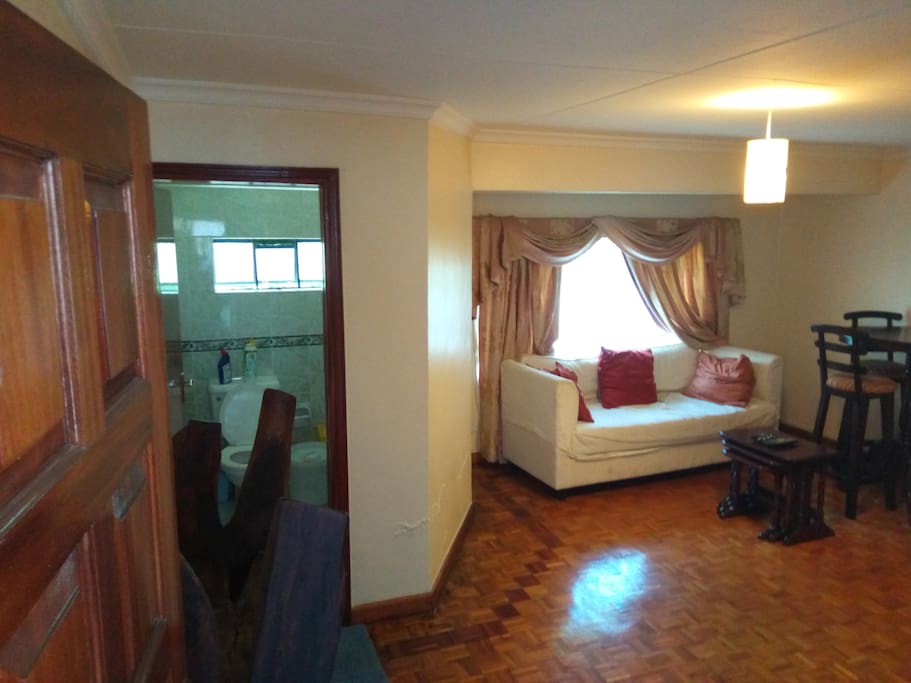 The view from the main entrance. Classy wooden multipurpose rack at the door, loo and sink for the visitors with a wash station included. The main sitting area with one 3 seater couch and 3 seater high table.