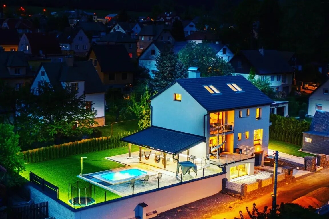 An exterior view of a modern house is presented at dusk, featuring a well-lit structure with large windows. A landscaped yard features a swimming pool surrounded by seating areas and hammocks. Neighboring houses are visible, with lights reflecting a cozy evening atmosphere.
