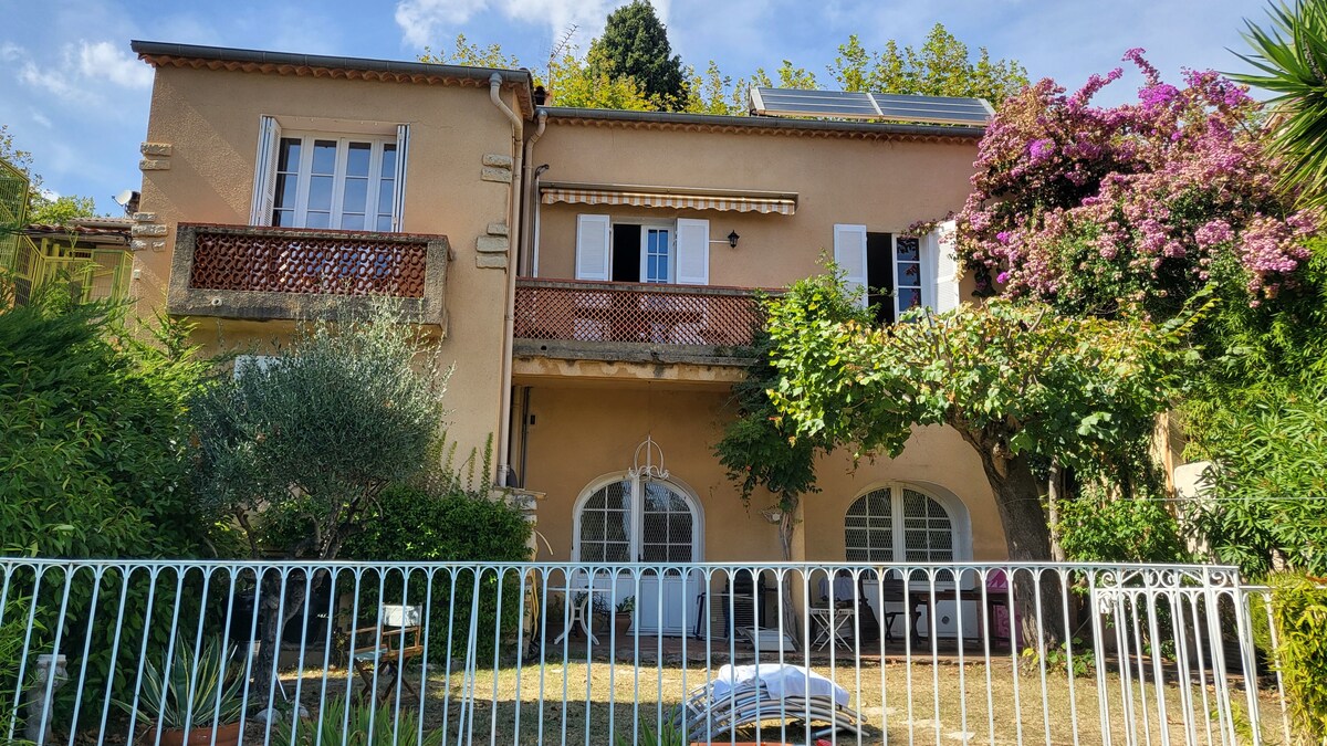 A charming townhouse is showcased, with a warm-toned exterior and decorative balcony. Lush greenery and flowering plants adorn the garden, while a white fence outlines the property. Sunlight filters through the trees, highlighting the inviting outdoor space.