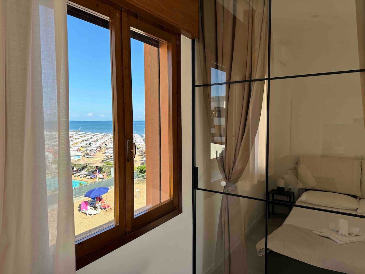 A large window offers a view of the beach and ocean, with colorful sun umbrellas visible on the sand. Sheer curtains frame the window, allowing natural light to fill the room, which features a glass partition separating the sleeping area.
