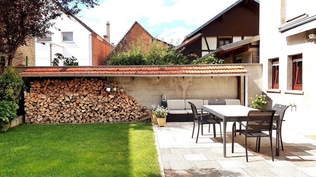 An outdoor space features a neatly organized woodpile beneath a tiled roof. An outdoor dining table with several chairs is present, surrounded by a well-maintained lawn and paved area. Nearby, potted plants add greenery to the setting, with a backdrop of houses and trees.