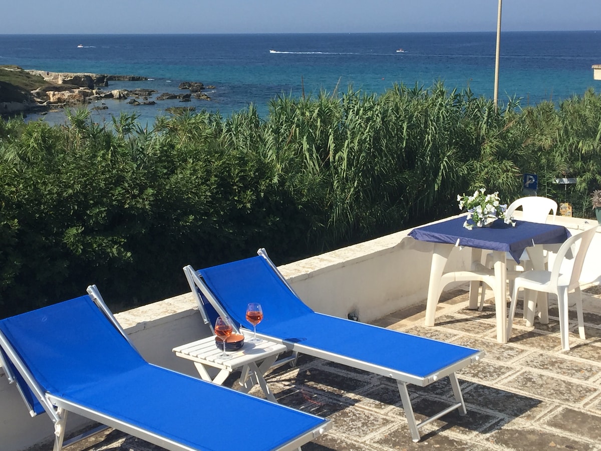A rooftop terrace is shown with two blue sun loungers positioned beside a small table. A glass of beverages rests on the table, accompanied by a floral arrangement. In the background, the expansive view of the sea and rocky coastline is visible, framed by coastal vegetation.