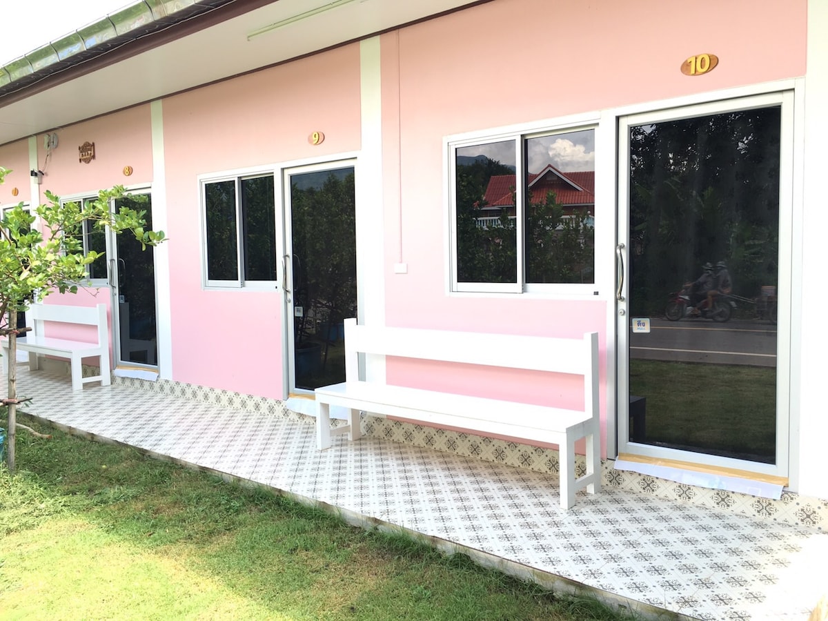 A row of rooms is presented in soft pink exterior, each featuring large windows and a door with a clear glass panel. White benches are positioned outside, providing seating options on the tiled walkway that extends across a grassy area.