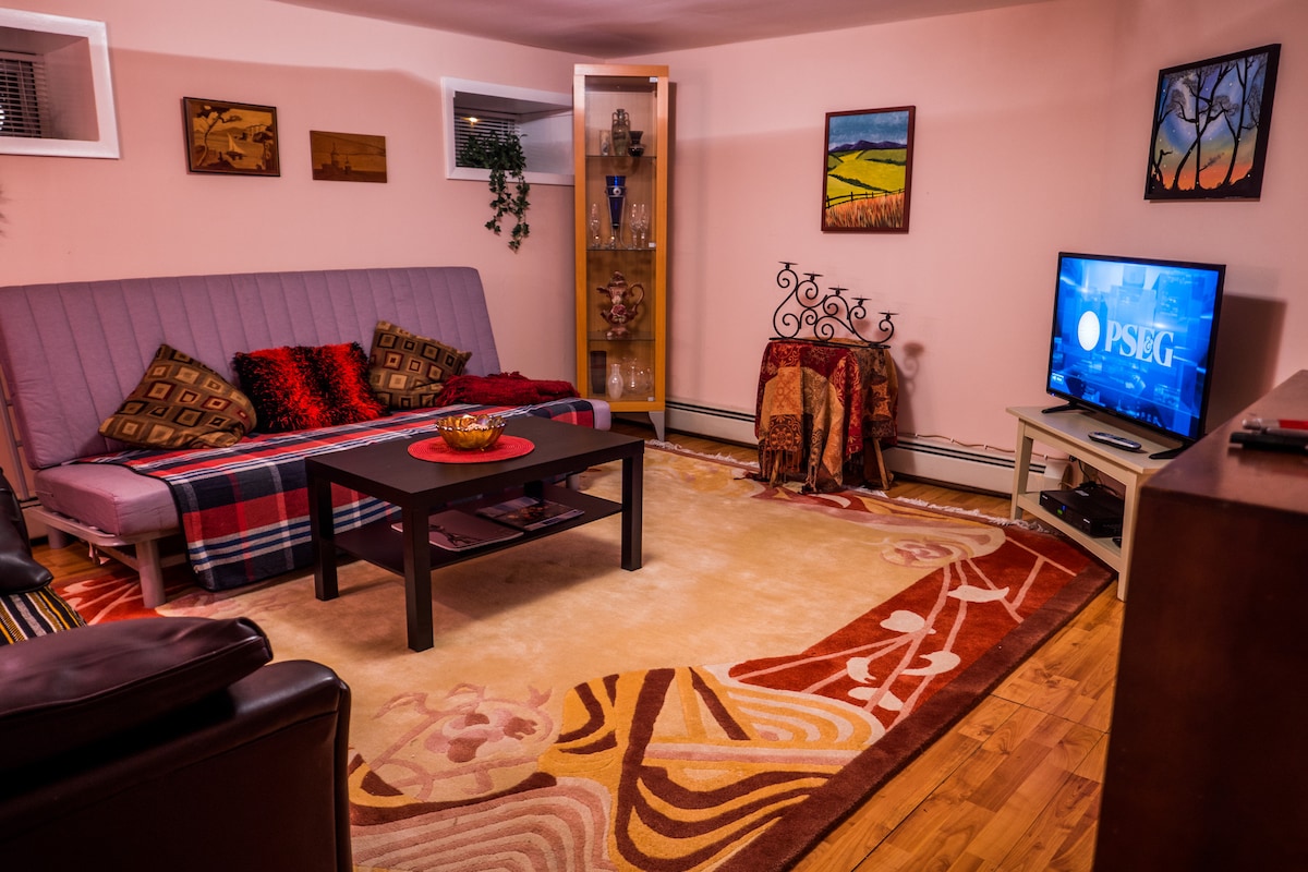 The living area features a futon with colorful blankets, complemented by accent pillows. A coffee table is situated at its center, surrounded by an area rug with floral patterns. A television is mounted on a unit, and wall art adds visual interest to the space.