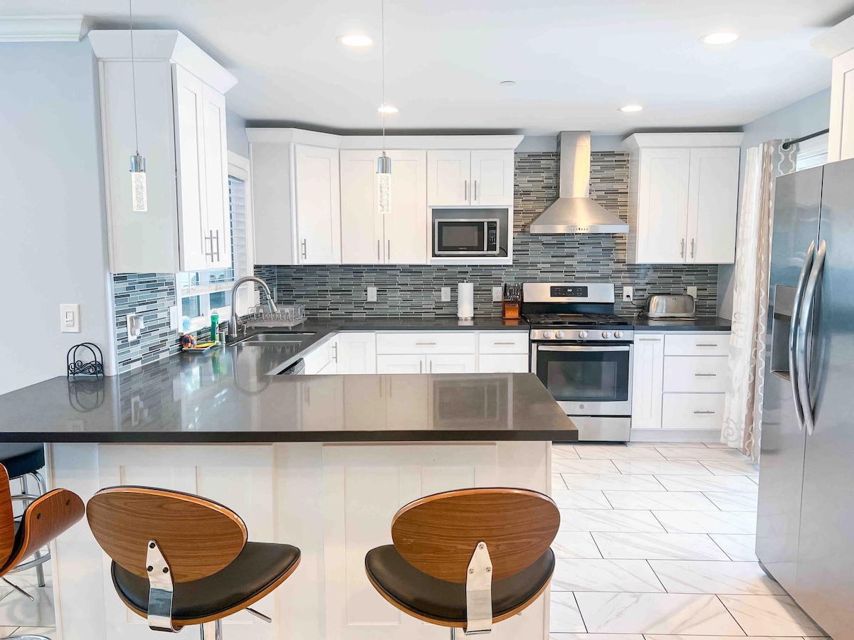 A modern kitchen is showcased with white cabinetry and a dark countertop. Stainless steel appliances, including a microwave and oven, are present. A mosaic backsplash adds texture. Three stools with wooden seats are positioned at the kitchen island, creating an inviting space for dining.