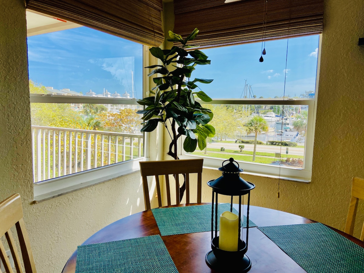 A dining area is captured with large windows providing views of the harbor. A wooden table is set with a textured mat and a lantern holding a candle. A potted plant adds greenery, enhancing the space's natural light.