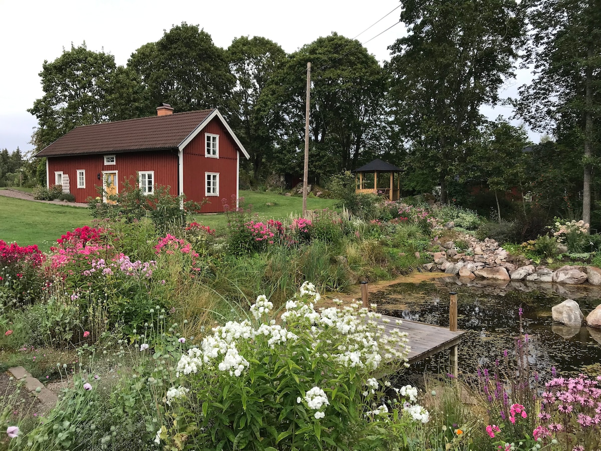 A charming red cabin is surrounded by vibrant flower beds, featuring various colorful blooms. A small pond with a wooden platform is visible nearby, complemented by natural rock formations. A gazebo sits in the garden, providing a peaceful spot within the lush greenery.
