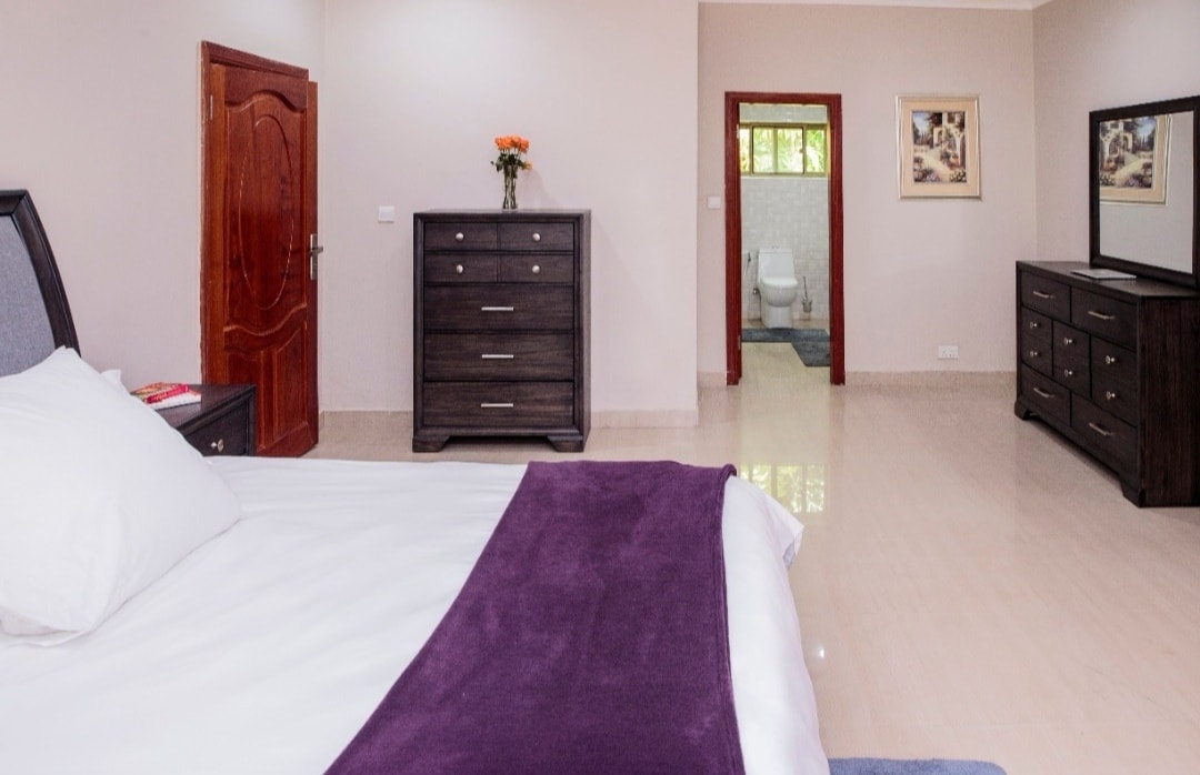 A spacious bedroom is furnished with a large bed covered by a silky purple throw. A dark wooden dresser stands nearby, complemented by a small vase of orange flowers. The room is well-lit, revealing an ensuite bathroom in the background.