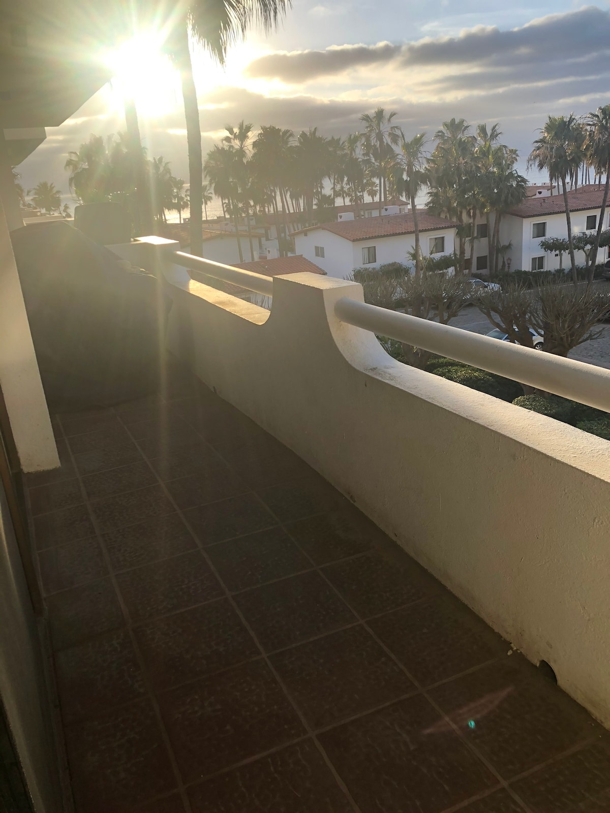 A balcony is visible, featuring a tiled floor and a railing overlooking palm trees and a neighboring building. Sunlight streams in from the side, creating a warm glow against the background of a cloudy sky.