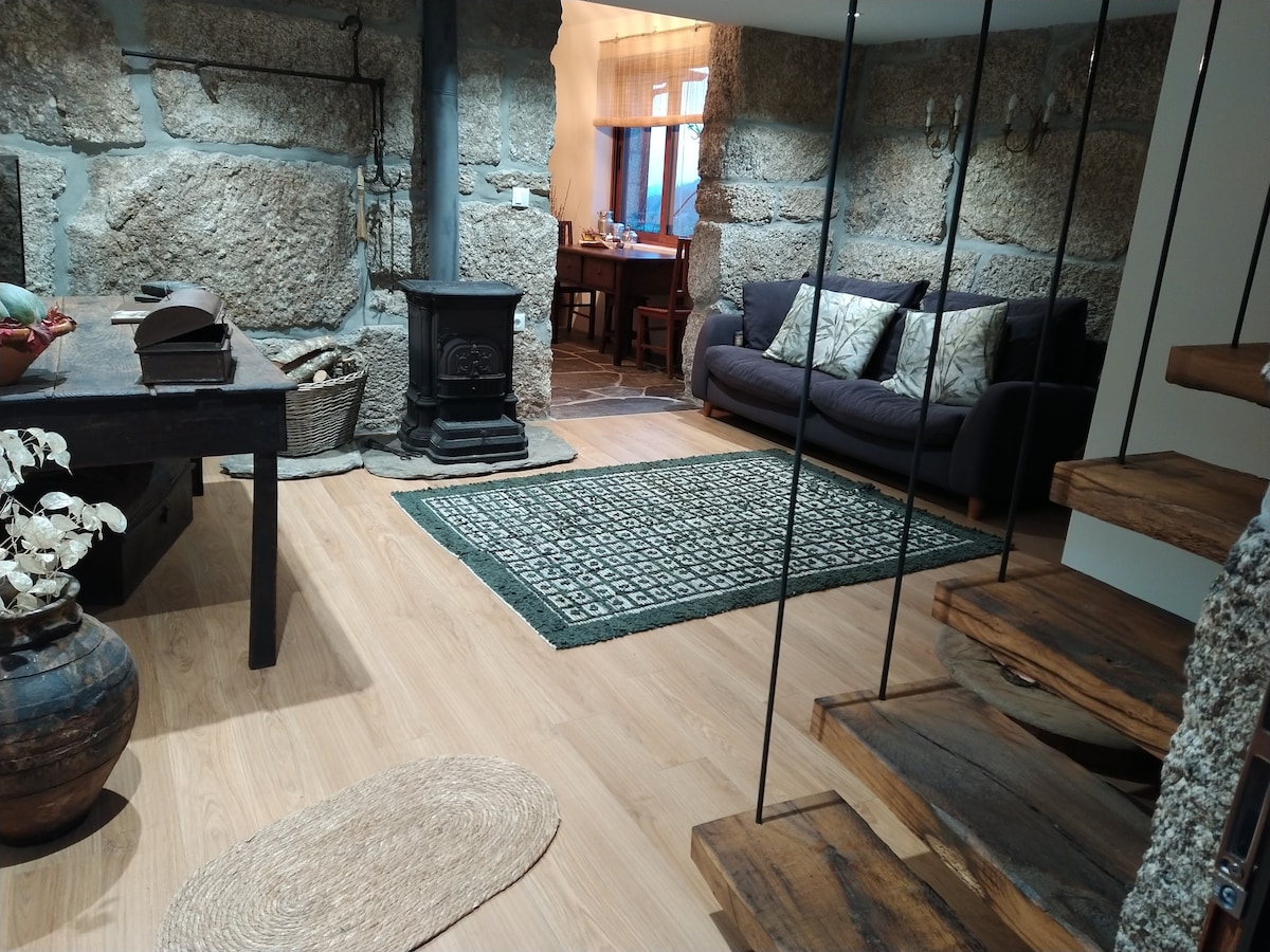 The living area features exposed stone walls and a wood stove, adding rustic charm. A cozy sofa is positioned near a dining table, with natural light illuminating the space through nearby windows. A woven rug and decorative elements enhance the inviting ambiance.