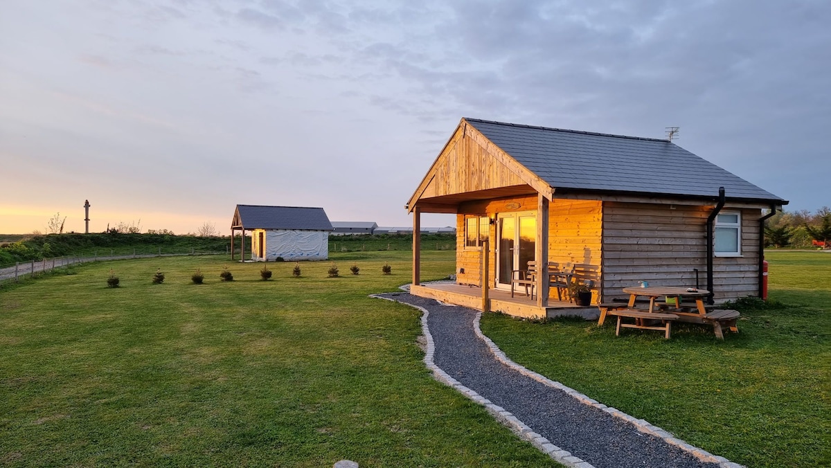 A wooden cabin with a sloped roof is set against a sunset sky, showcasing large glass doors that offer a view of the outdoors. A pathway made of gravel leads to the entrance, surrounded by well-maintained grass and landscaping, with another cabin visible in the background.