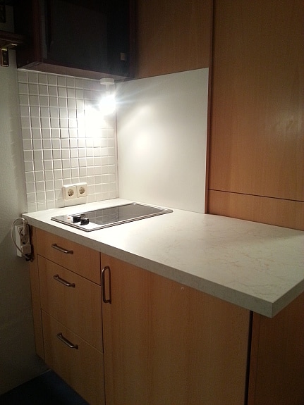 A compact kitchen area is shown, featuring a smooth countertop with a built-in stove. Wooden cabinetry provides storage, and a small backsplash of square tiles lines the wall. Soft lighting from above illuminates the workspace, creating a functional cooking environment.