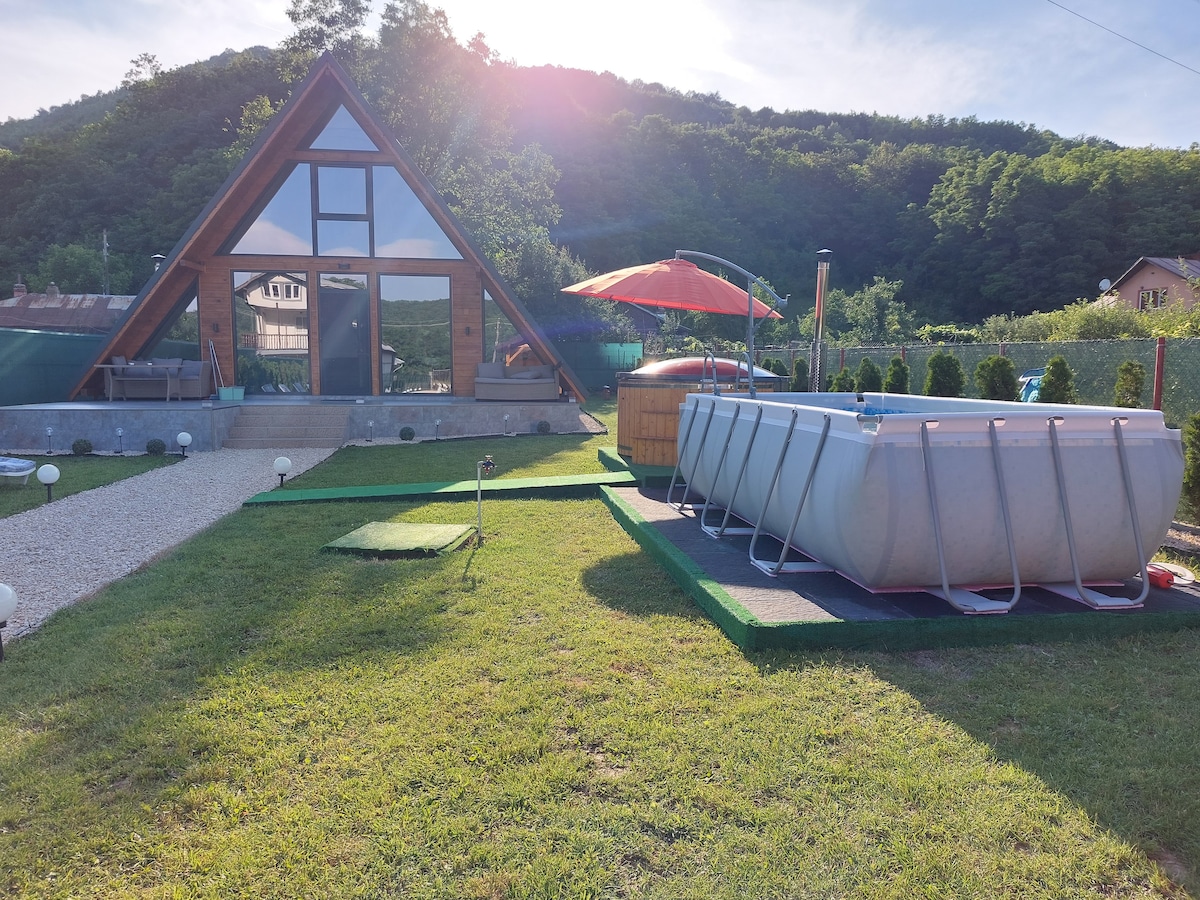 A spacious A-frame cabin is set against a backdrop of lush green hills. An above-ground pool is positioned beside the cabin, with a sun patio featuring a large umbrella. Well-maintained grass and gravel paths lead to the cabin's entrance.