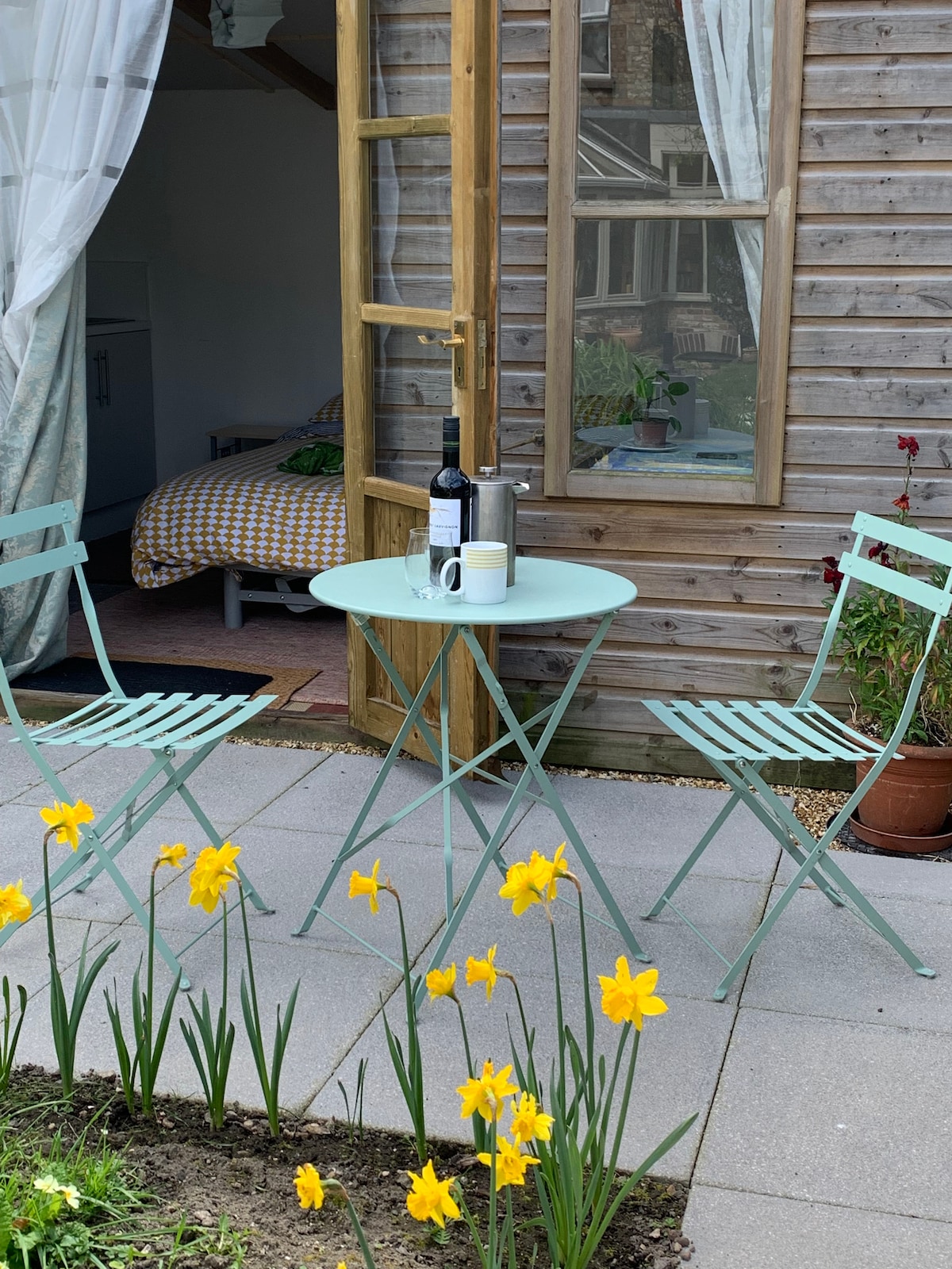 A patio area features a small round table with two green chairs, surrounded by blooming yellow daffodils. The entrance to the shed is visible in the background, with a patterned bedspread visible inside. A bottle and two glasses are set on the table.