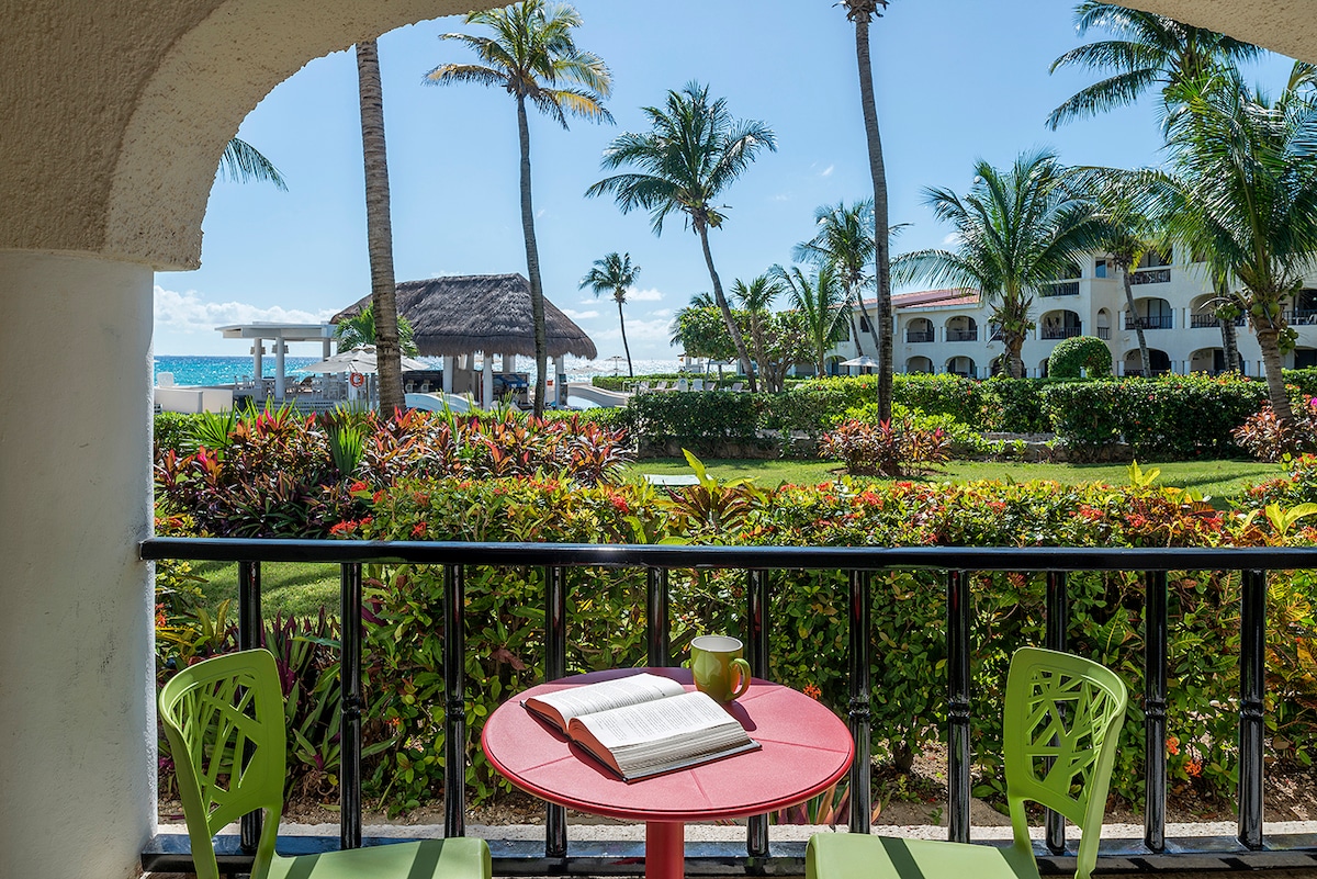 A small outdoor balcony is shown, featuring a round red table and two green chairs. A book rests open on the table, with vibrant greenery and palm trees visible in the background. The Caribbean Sea can be seen beyond the lush landscape.