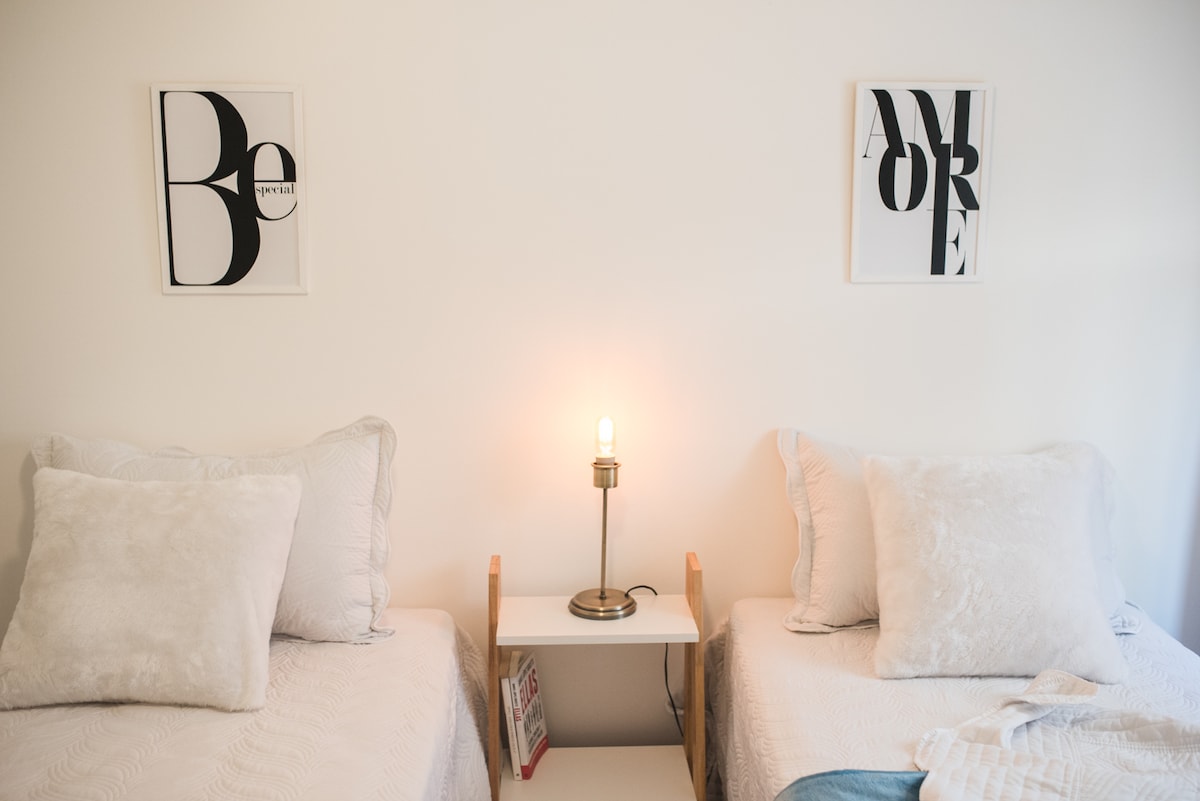 Two individual beds are positioned side by side, each adorned with light-colored linens and decorative pillows. A small nightstand is placed between them, featuring a stylish lamp that provides a soft glow. Above each bed, framed art prints add a subtle artistic touch to the room.