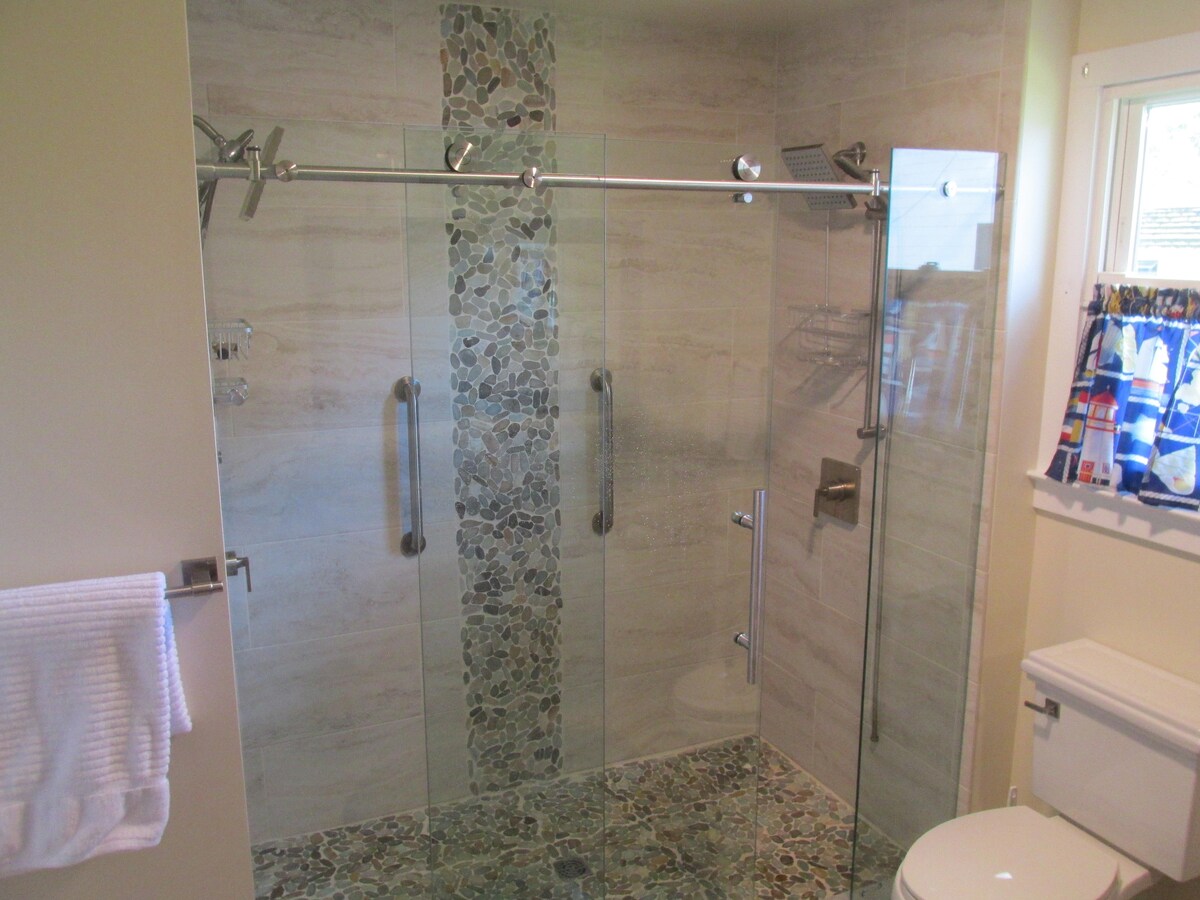 A spacious bathroom features a walk-in shower enclosed by glass. The shower includes a decorative stone accent and chrome fixtures. Neutral tones dominate the space with a contemporary tile design on the floor. A white towel hangs neatly on a rack, and natural light enters through a nearby window.