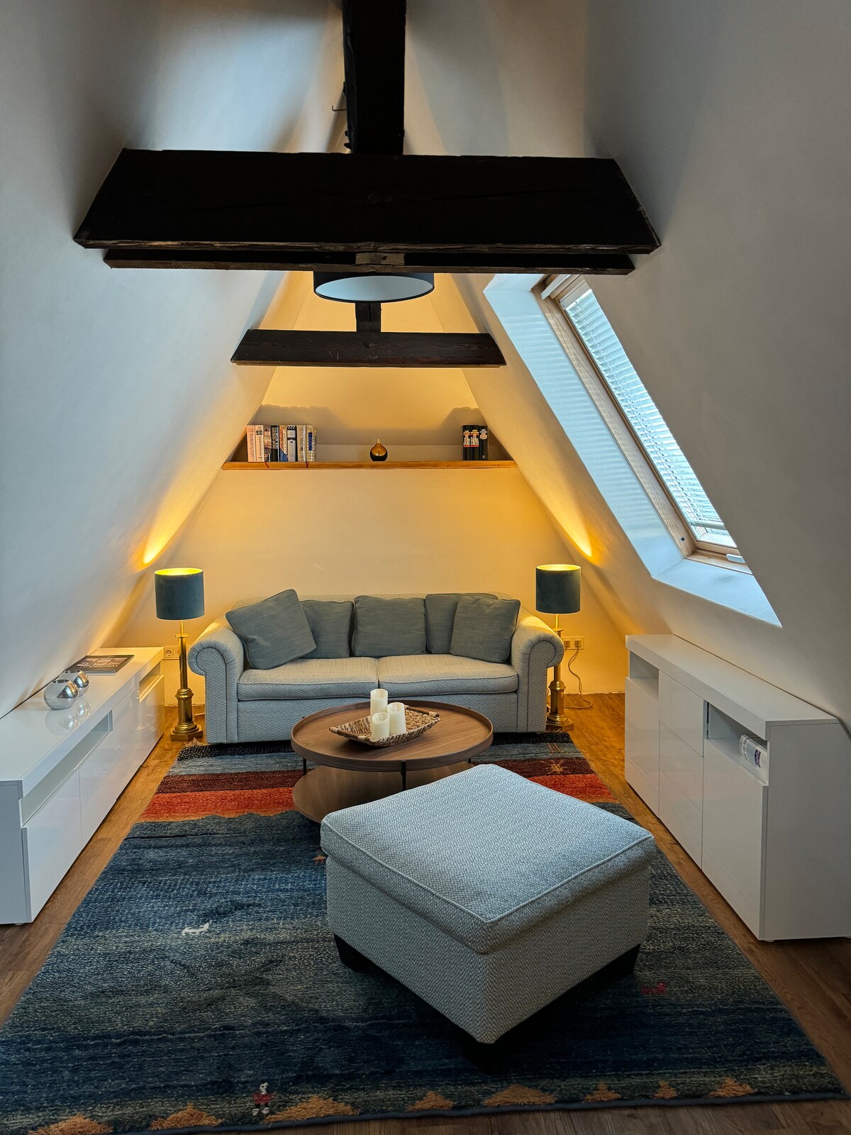 A cozy living area is presented with a light-colored sofa and a round coffee table at the center. Blue lamps provide soft illumination alongside a patterned area rug. Angled roof beams and a small window with shutters create a unique architectural feature.