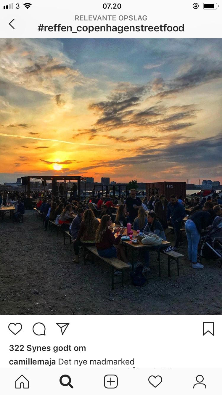 A lively dining scene is set outdoors as people gather around long wooden tables, enjoying food during sunset. The sun casts a warm glow, with scattered clouds enhancing the evening sky. Food stalls and a vibrant atmosphere reflect a bustling street food market.