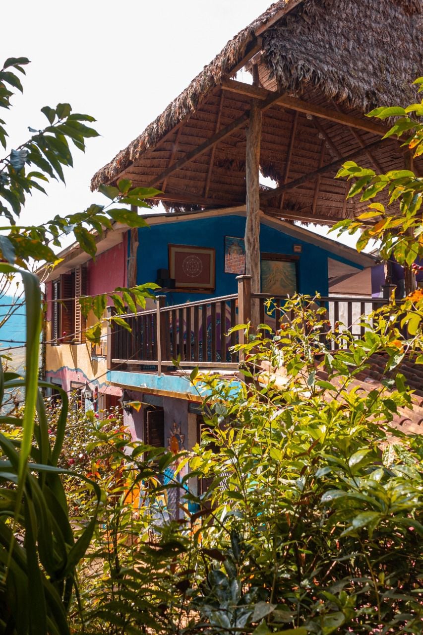 A rustic structure with a thatched roof is surrounded by vibrant greenery. Colorful walls are visible, enhancing the natural setting. A balcony extends from the upper level, offering an outdoor space to enjoy the surrounding landscape. The overall ambiance reflects a harmonious blend with nature.