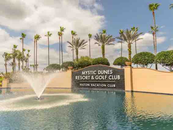 A welcoming entrance to Mystic Dunes Resort & Golf Club is featured, showcasing a prominent sign set against a tranquil water feature. Palm trees are positioned along the perimeter, contributing to the resort's tropical ambiance under a partly cloudy sky.